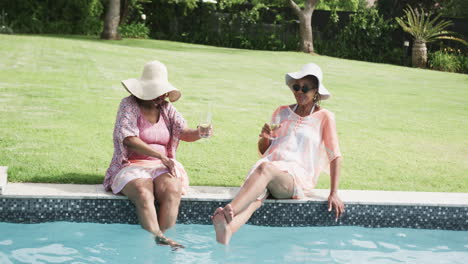 Glückliche-ältere-Afroamerikanische-Freundinnen-In-Sonnenhüten,-Die-Mit-Getränken-Am-Pool-Sitzen,-Zeitlupe