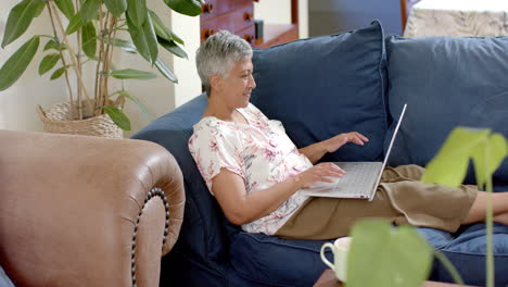 Happy-senior-biracial-woman-sitting-on-couch-and-using-laptop-at-home,-slow-motion