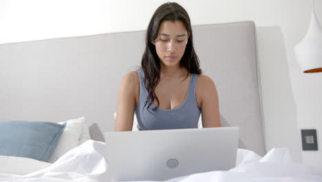 Focused-biracial-teenage-girl-sitting-on-bed-in-the-morning-using-laptop,-copy-space,-slow-motion