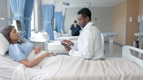 Diverse-female-patient-in-hospital-bed-and-male-doctor-with-tablet-talking-in-ward,-slow-motion
