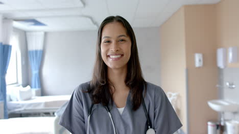 Portrait-of-smiling-biracial-female-doctor-with-long-dark-hair-in-hospital-ward,-slow-motion