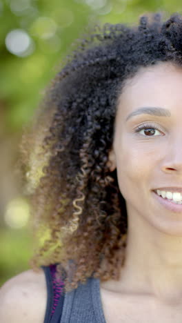 Vertical-video-half-portrait-of-biracial-woman-with-long-hair-smiling-in-sunny-garden,-slow-motion