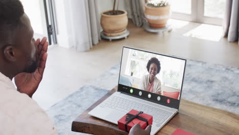 African-american-man-holding-red-gift-and-using-laptop-with-african-american-woman-on-screen