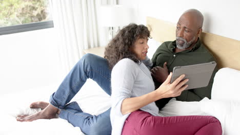 Happy-mature-diverse-couple-sitting-on-bed-using-tablet-and-talking-at-home,-slow-motion
