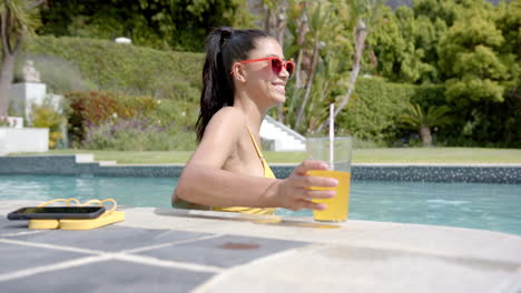 Happy-biracial-teenage-girl-in-sunglasses-standing-in-sunny-pool-with-drink,-slow-motion