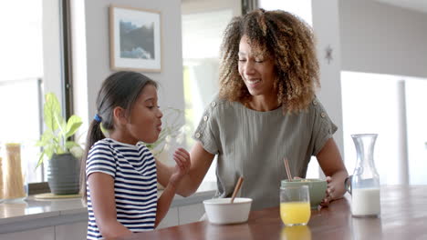 Happy-biracial-mother-and-daughter-eating-breakfast-cereal-in-sunny-kitchen,-slow-motion