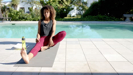 Happy-biracial-woman-practicing-yoga-sitting-by-pool-in-sunny-garden,-copy-space,-slow-motion