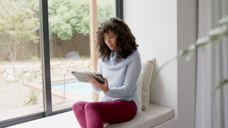 Happy-mature-caucasian-woman-sitting-at-window-and-using-tablet-at-home,-slow-motion