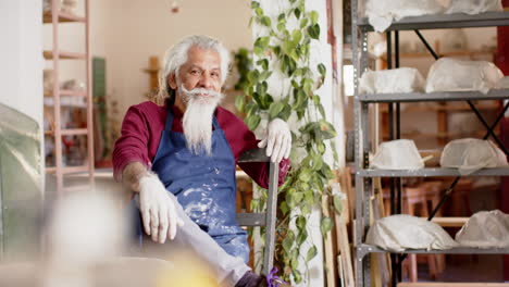 Happy-senior-biracial-potter-with-long-beard-resting-and-smiling-in-pottery-studio,-slow-motion