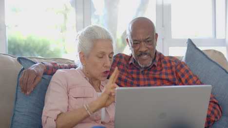 Glückliches-älteres,-Gemischtrassiges-Paar,-Das-Zu-Hause-Auf-Der-Couch-Sitzt-Und-Einen-Laptop-Benutzt,-Zeitlupe