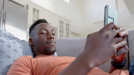 Happy-african-american-man-lying-and-using-smartphone-in-sunny-living-room,-slow-motion