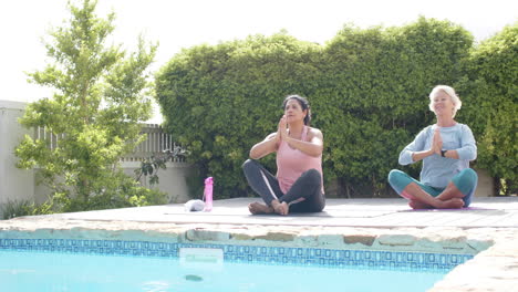 Dos-Mujeres-Mayores-Felices-Y-Diversas-Practicando-Yoga-Junto-A-La-Piscina-En-Un-Jardín-Soleado,-Cámara-Lenta,-Espacio-Para-Copiar