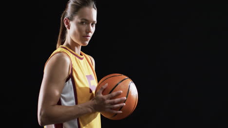 Confident-young-Caucasian-woman-holds-a-basketball-on-a-black-background,-with-copy-space