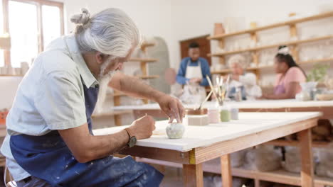 Focused-biracial-potter-with-long-beard-glazing-clay-jug-in-pottery-studio,-slow-motion