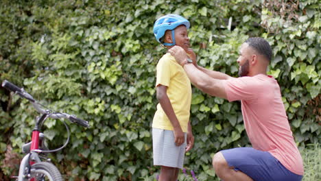 Happy-african-american-father-putting-cycle-helmet-on-son-in-garden,-copy-space,-slow-motion