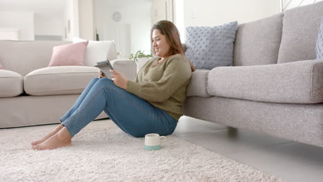 Happy-plus-size-biracial-woman-relaxing-in-living-room-using-tablet,-copy-space,-slow-motion