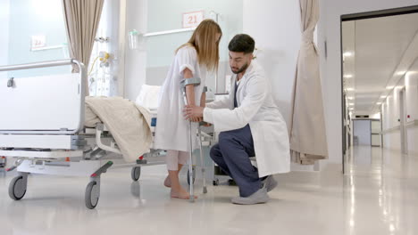 Diverse-male-doctor-helping-girl-patient-use-crutches-in-hospital-ward,-slow-motion