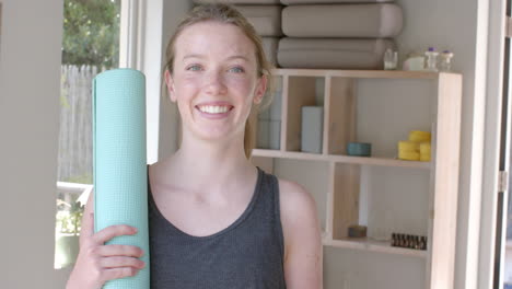 Portrait-of-happy-caucasian-fitness-teenage-girl-holding-yoga-mat-in-white-room,-slow-motion