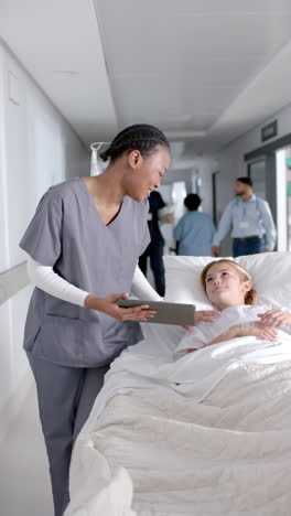 Vertical-video-of-diverse-doctor-and-girl-patient-lying-on-bed-using-tablet-in-hospital,-slow-motion