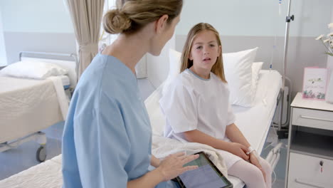 Caucasian-female-doctor-using-tablet-talking-with-girl-patient-sitting-on-hospital-bed,-slow-motion