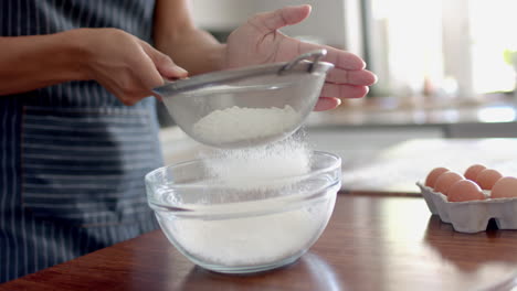 Midsection-of-biracial-woman-in-apron-sieving-flour,-baking-in-kitchen,-slow-motion
