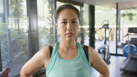 Serious-asian-woman-with-arms-akimbo-looking-at-camera-in-sunny-home-gym,-slow-motion