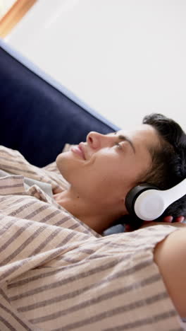 Vertical-video-of-biracial-man-in-headphones-lying-on-couch,-slow-motion