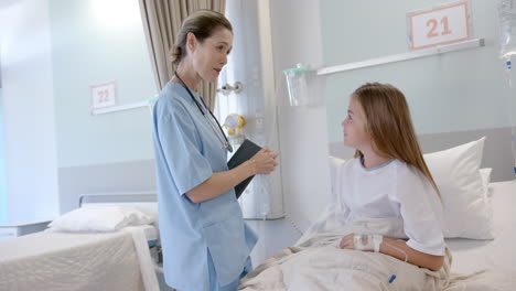 Happy-caucasian-female-doctor-with-tablet-talking-with-girl-patient-in-hospital-bed,-slow-motion