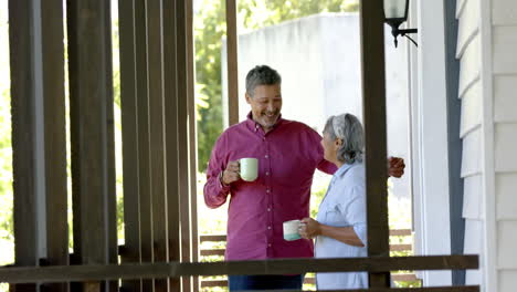 Happy-biracial-senior-couple-embracing-at-window,-talking-and-drinking-tea