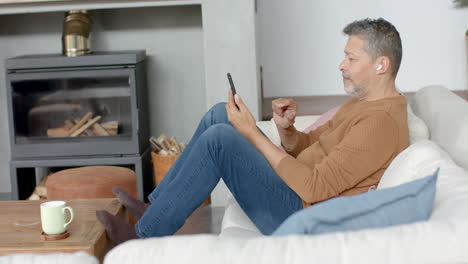 Älterer-Mann-Mit-Gemischter-Abstammung,-Der-Smartphone-Und-Kopfhörer-Benutzt-Und-Auf-Der-Couch-Im-Wohnzimmer-Sitzt,-Zeitlupe