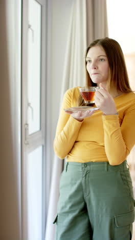 Vertical-video-of-thoughtful-caucasian-woman-drinking-tea-looking-through-window,-slow-motion