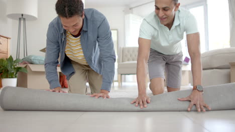 Happy-diverse-gay-male-couple-moving-into-new-home,-unrolling-rug-in-living-room,-slow-motion