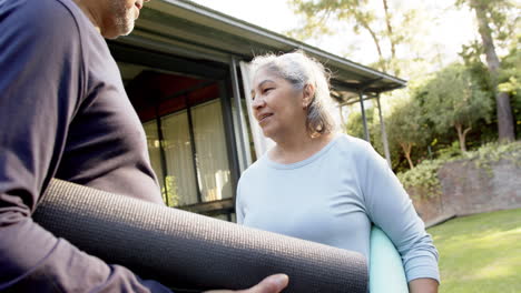 Happy-diverse-senior-couple-talking-and-holding-exercise-mat-in-garden