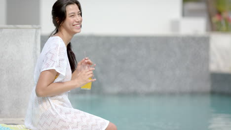 Happy-biracial-teenage-girl-holding-juice-sitting-by-pool,-copy-space,-slow-motion