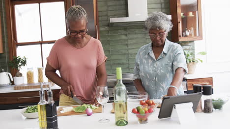 Senior-african-american-female-friends-cooking,-chopping-vegetables-and-using-tablet,-slow-motion