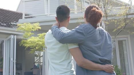 Rear-view-of-happy-diverse-gay-male-couple-embracing-in-sunny-garden-of-new-home,-slow-motion