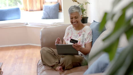 Glückliche-ältere-Frau-Mit-Gemischter-Abstammung-Auf-Der-Couch,-Die-Eine-Kreditkarte-Hält-Und-Zu-Hause-Ein-Tablet-Benutzt,-Zeitlupe