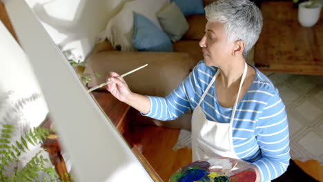 Mujer-Birracial-Senior-Enfocada-Usando-Delantal-Y-Pintando-Sobre-Lienzo-Grande-En-Casa,-Cámara-Lenta