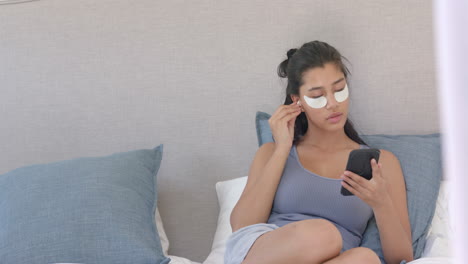 Biracial-teenage-girl-wearing-under-eye-masks-sitting-bed-using-smartphone,-copy-space,-slow-motion