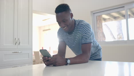 Glücklicher-Afroamerikanischer-Mann,-Der-Sich-Auf-Die-Theke-Lehnt-Und-Sein-Smartphone-In-Einer-Sonnigen-Küche-Benutzt,-Zeitlupe