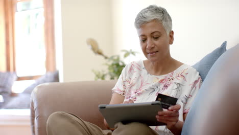 Happy-senior-biracial-woman-on-couch-holding-credit-card-and-using-tablet-at-home,-slow-motion