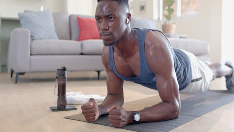 Tired-african-american-man-doing-plank,-using-headphones-and-smartphone-in-living-room,-slow-motion