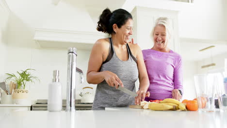 Two-happy-diverse-senior-women-chopping-fruits-and-laughing-in-sunny-kitchen,-slow-motion
