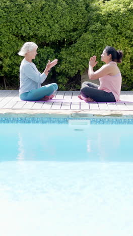 Two-happy-diverse-senior-women-doing-yoga-meditation-by-pool-in-sunny-garden,-slow-motion