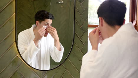 Focused-biracial-man-applying-under-eye-masks-looking-in-bathroom-mirror,-slow-motion