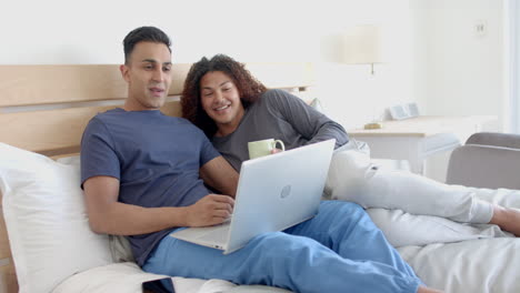 Happy-diverse-gay-male-couple-on-bed,-having-coffee-and-using-laptop-in-the-morning,-slow-motion