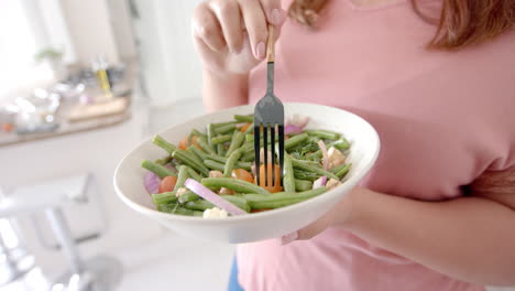 Midsection-of-plus-size-biracial-woman-eating-vegetable-salad-standing-in-kitchen,-slow-motion
