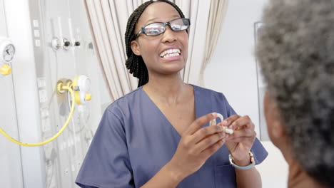 African-american-female-doctor-applying-hearing-aid-to-senior-woman-in-hospital-room,-slow-motion
