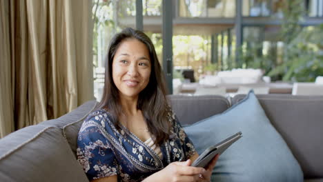 Happy-asian-woman-using-tablet-and-looking-out-window-in-sunny-living-room,-slow-motion