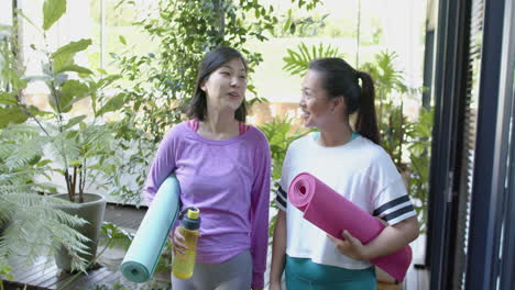 Glückliche-Asiatische-Freundinnen-Mit-Trainingsmatten-Gehen-Und-Lächeln-Auf-Der-Sonnigen-Terrasse,-Zeitlupe